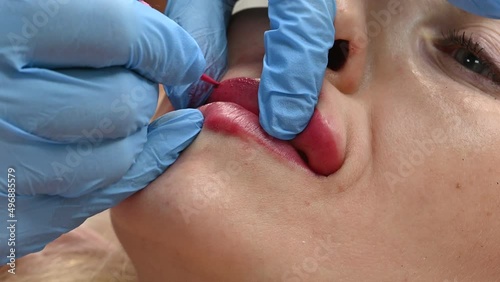 Skilled cosmetologist applying permanent makeup on lips in a beauty salon. Woman undergoing procedure of lip blushing with tattoo machine. Close-up