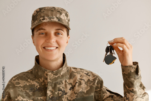 Portrait of smiling positive optimistic military woman wearing camouflage uniform and cap, standing looking at camera with happy smile and holding keys from a new car or apartment.