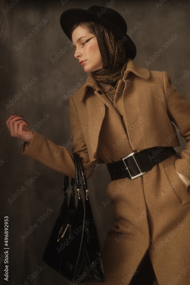 A beautiful young woman poses in the studio on a gray background. Vintage look. Autumn or spring coat, black hat and scarf. Large eye arrows.