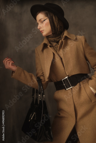 A beautiful young woman poses in the studio on a gray background. Vintage look. Autumn or spring coat, black hat and scarf. Large eye arrows.