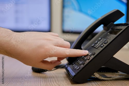 A man hand dials a number on the phone at the monitors of office computers. Finger presses on the buttons of a office landline phone
