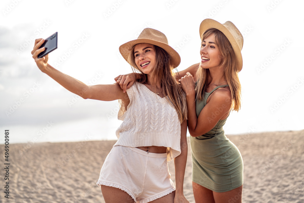 Two happy blonde caucasian sisters having fun on the beach during summer, taking selfie with mobile phone, smiling and looking to the smartphone camera. Beautiful girls wearing hats. Friendship.
