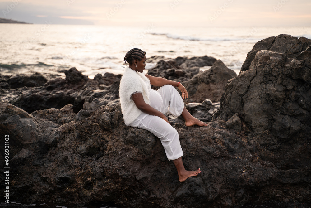 Beautiful pregnant woman in white clothes posing on the rocky coast, sitting and relaxing during sunset. Future motherhood concept. Mom to be.