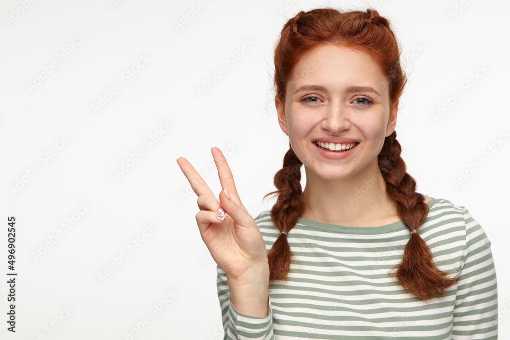 Indoor portrait of young ginger female standing over white background showing v sign into camera Indoor portrait of young ginger female standing over white background showing v sign into camera