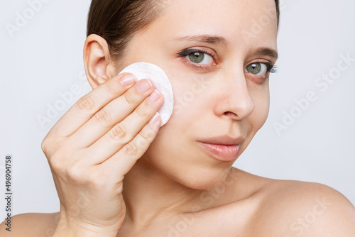 Close up of young caucasian woman cleaning her face with lotion using cotton pad isolated on a white background. Skin care, cosmetology. Evening routine. The girl washes off her makeup with a cleanser
