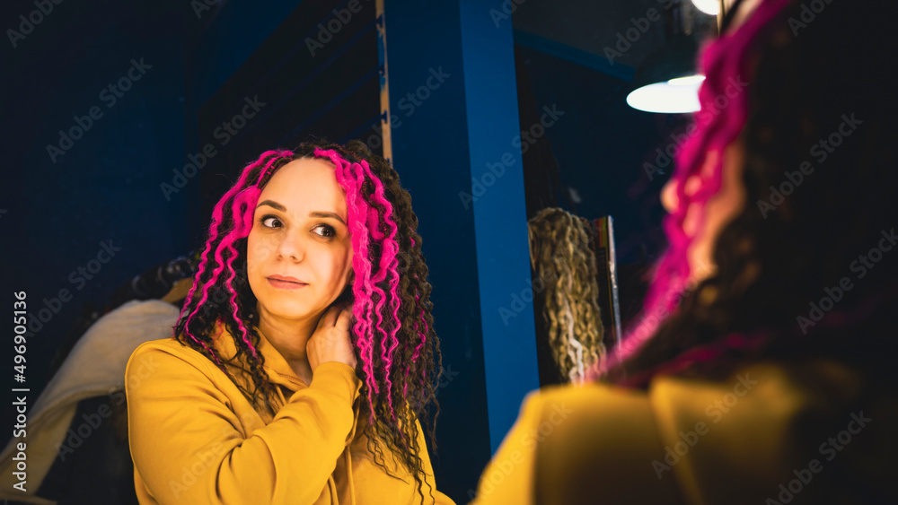 Fototapeta premium Young woman looking in mirror, considering her new hairstyle. Beautiful female with dreadlocks preening in front of mirror.