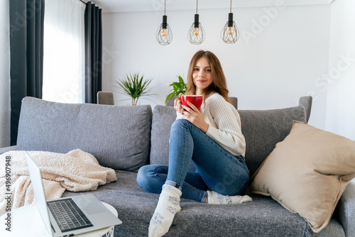 Young beautiful girl is sitting on the sofa with a cup of tea or coffee and a laptop