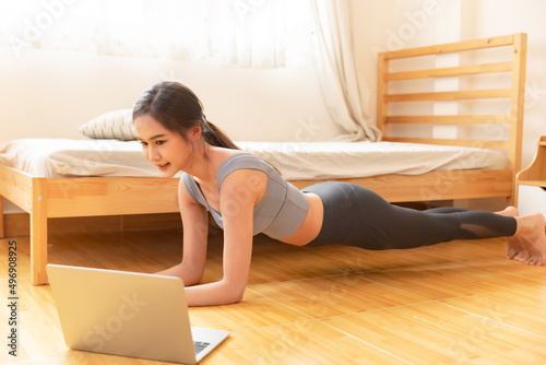 Young Asian woman is exercising yoga at home. Fitness, workout, healthy living and diet concept. Female watching videos online on laptop computer in bedroom.