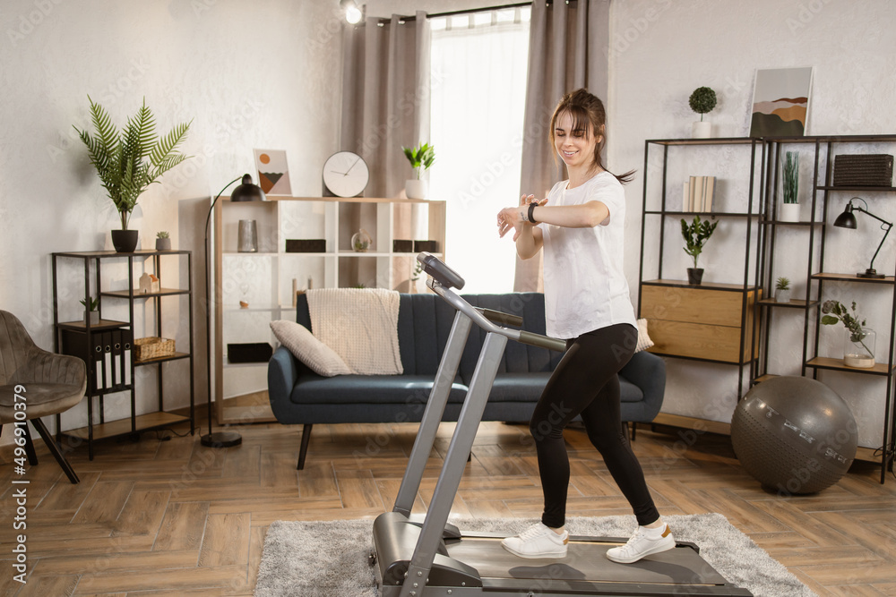 Beautiful caucasian women with long black and tied hair in a sports leggings and a white T-shirt and sneakers doing exercise by running on the electric treadmill in the fitness room .