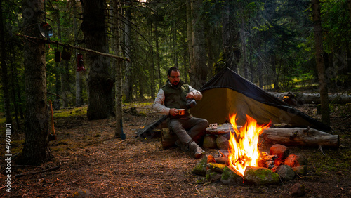 camper man sitting by campfire in spring forest and drinking tea