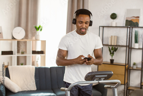 Jogging workout. Joyful african man in modern headphones chooses music for listening while running on treadmill at home. Motivated african american guy in sportswear spending time at gym, jogging