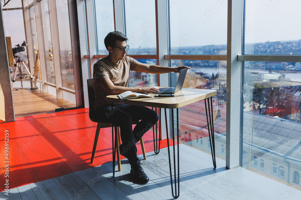 Young handsome smiling smart freelancer in glasses works at a laptop while spending time in a cafe. A positive student sits at a wooden table in a cafe. Happy man sitting while working indoors Young handsome smiling smart freelancer in glasses works at a laptop while spending time in a cafe. A positive student sits at a wooden table in a cafe. Happy man sitting while working indoors