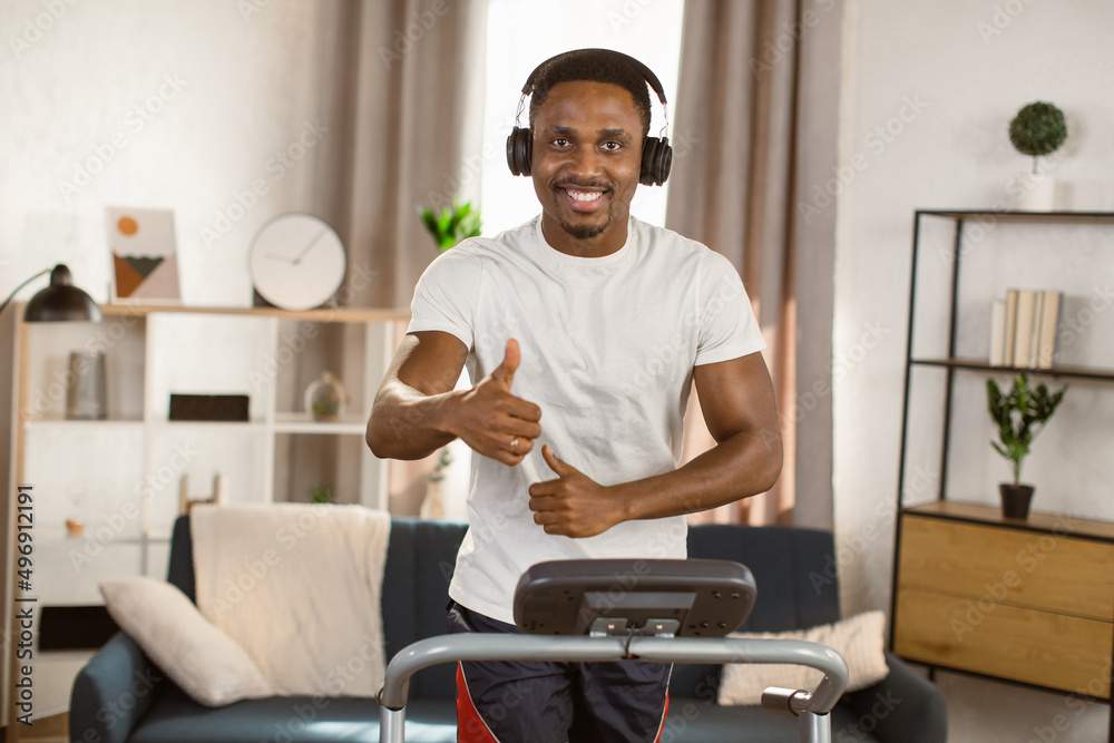 Young athletic african american man running on treadmill at home. Handsome muscular guy working out and listening to music with headphones looking at the camera. Enjoy your lifestyle.