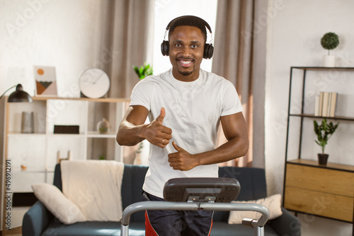 Young athletic african american man running on treadmill at home. Handsome muscular guy working out and listening to music with headphones looking at the camera. Enjoy your lifestyle.