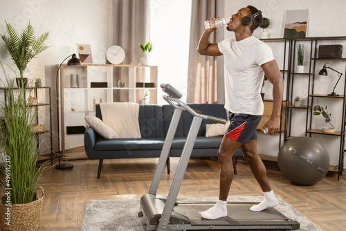 Happy young african american male athlete in headphones drinking bottled water enjoying favorite song while running on treadmill during intensive workout at modern apartment.