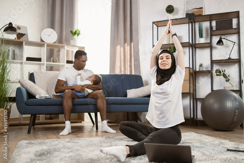 Arabic girl in hijab practicing online yoga  watching video tutorial on laptop  excersising in living room. African American man putting little son to sleep while wife going in for sports on weekend