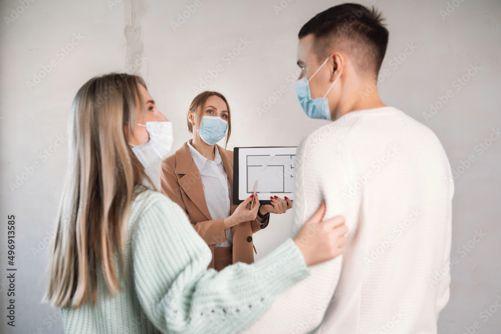 Fototapeta premium Woman in suit talking to couple and show apartment plan in unfinished building. All people in masks. Life in pandemic