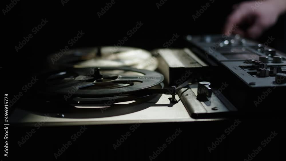 close up old reel-to-reel tape recorder plays magnetic tape record. Hand push start button. Beautiful gloomy shot of vintage retro turntable on table dark light. beam of light falls on old reel player