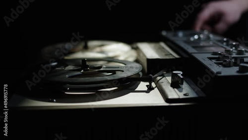 close up old reel-to-reel tape recorder plays magnetic tape record. Hand push start button. Beautiful gloomy shot of vintage retro turntable on table dark light. beam of light falls on old reel player