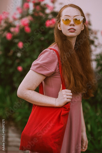 Young beautiful red-haired long-haired woman in a summer pink dress walks through the streets of a summer city.