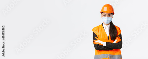 Covid-19 safety protocol at enterpise, construction and preventing virus concept. Confident asian female chief engineer looking how workers follow protocol during coronavirus, wear face mask photo