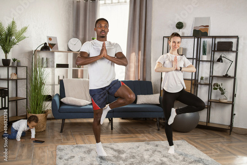 Loving caucasian woman with her african american husband doing fitness training while their mixed race son is playing on the floor at home. Doing yoga together. Healthy family lifestyle concept.