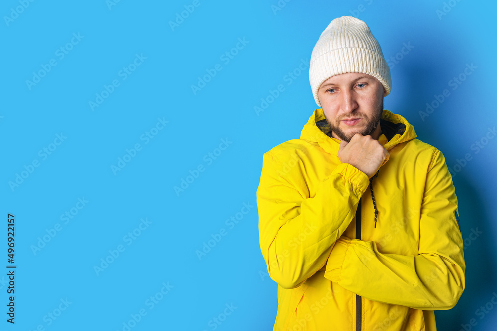 Pensive young man in a yellow jacket holding his hand at his chin on a blue background Pensive young man in a yellow jacket holding his hand at his chin on a blue background