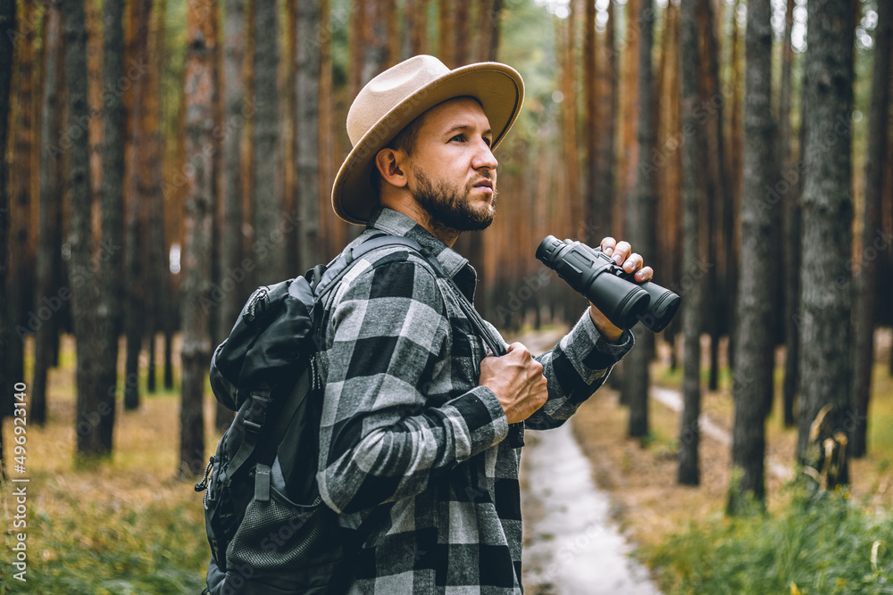 Man in a hat and a plaid shirt holds binoculars and walks through the forest