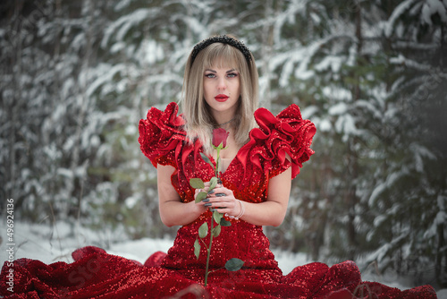 Young woman in the red dress in the snowy winter forest with red rose flower.