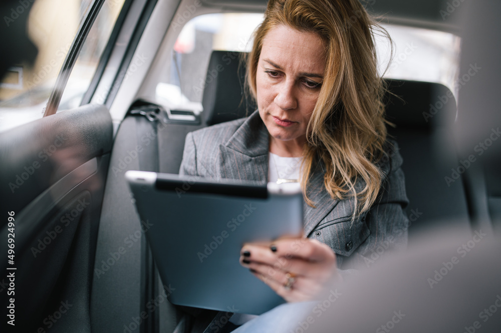 A business woman uses a tablet in the back seat of a car.