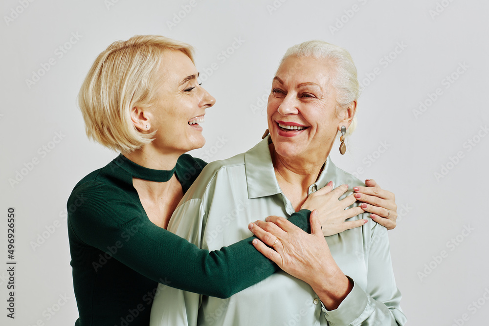 Candid waist up portrait of senior woman with adult daughter embracing and laughing happily in studio looking at each other Candid waist up portrait of senior woman with adult daughter embracing and laughing happily in studio looking at each other