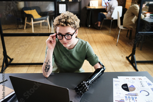 Girl in eyeglasses with prosthetic arm sitting at table with documents and looking at monitor of laptop at office