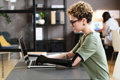Young woman with prosthetic arm typing on laptop while sitting at office desk Fototapeta