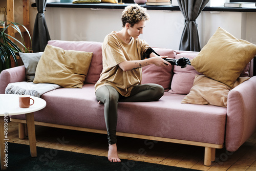Young woman sitting on sofa in living room and putting on prosthetic arm