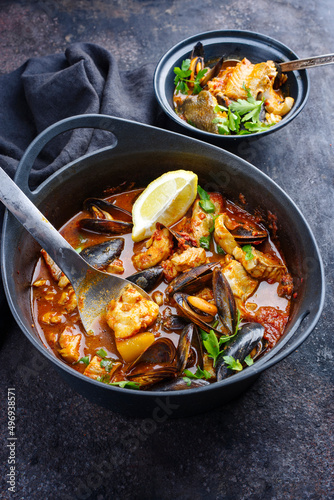 Modern style traditional Spanish seafood zarzuela de pescado with fish, king prawns and blue mussels served in red sauce as close-up in design pot