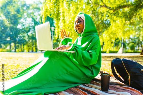 hispanic woman in green muslim hijab with bright make up and nose piercing working by laptop sit on grass outdoors summer park