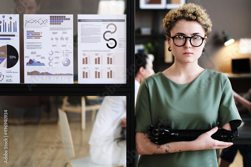 Portrait of young businesswoman with with prosthetic arm leaning on glass wall with financial reports and looking at camera