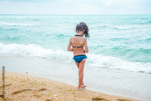 A little girl with curly hair in a blue bathing suit stands with her back to the camera on a sandy beach near the sea, ocean. Sea holidays, travel and beach holidays with children