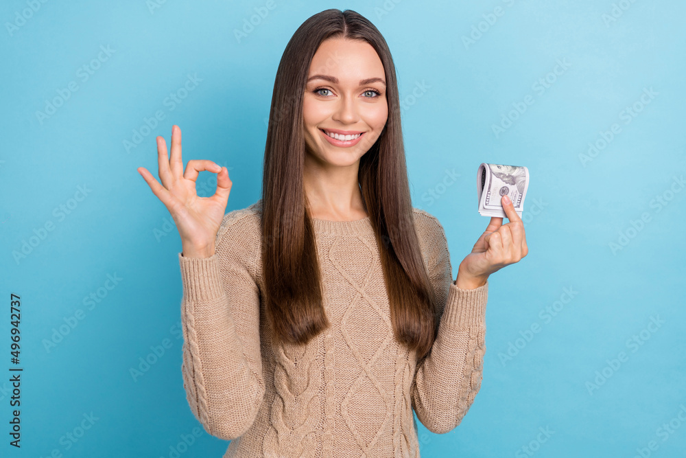Photo of cute young brunette lady hold money show okey wear brown sweater isolated on blue color background Photo of cute young brunette lady hold money show okey wear brown sweater isolated on blue color background