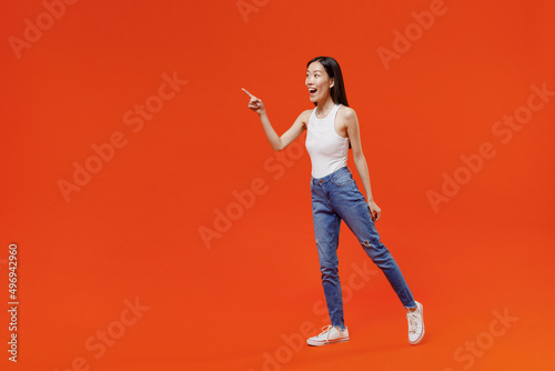 Full size body length side view impressed fun young woman of Asian ethnicity 20s in white tank top pointing index finger aside keep mouth wide open isolated on plain orange background studio portrait.