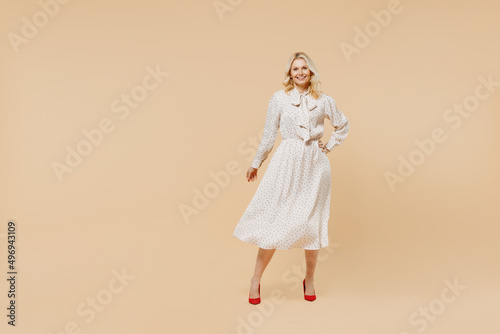 Full size body length blithesome elderly gray-haired blonde woman lady 40s years old wear pink dress hold hand on waist looking camera posing isolated on plain pastel beige background studio portrait