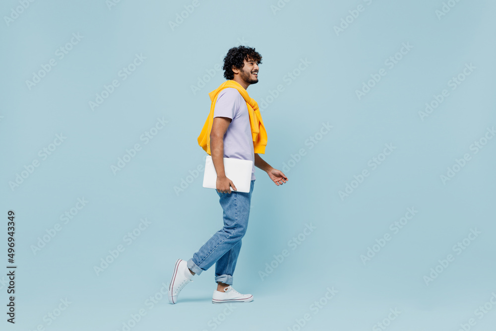 Full size body length side view exultant young bearded Indian man 20s wears white t-shirt hold use work on laptop pc computer stepping isolated on plain pastel light blue background studio portrait. Full size body length side view exultant young bearded Indian man 20s wears white t-shirt hold use work on laptop pc computer stepping isolated on plain pastel light blue background studio portrait.
