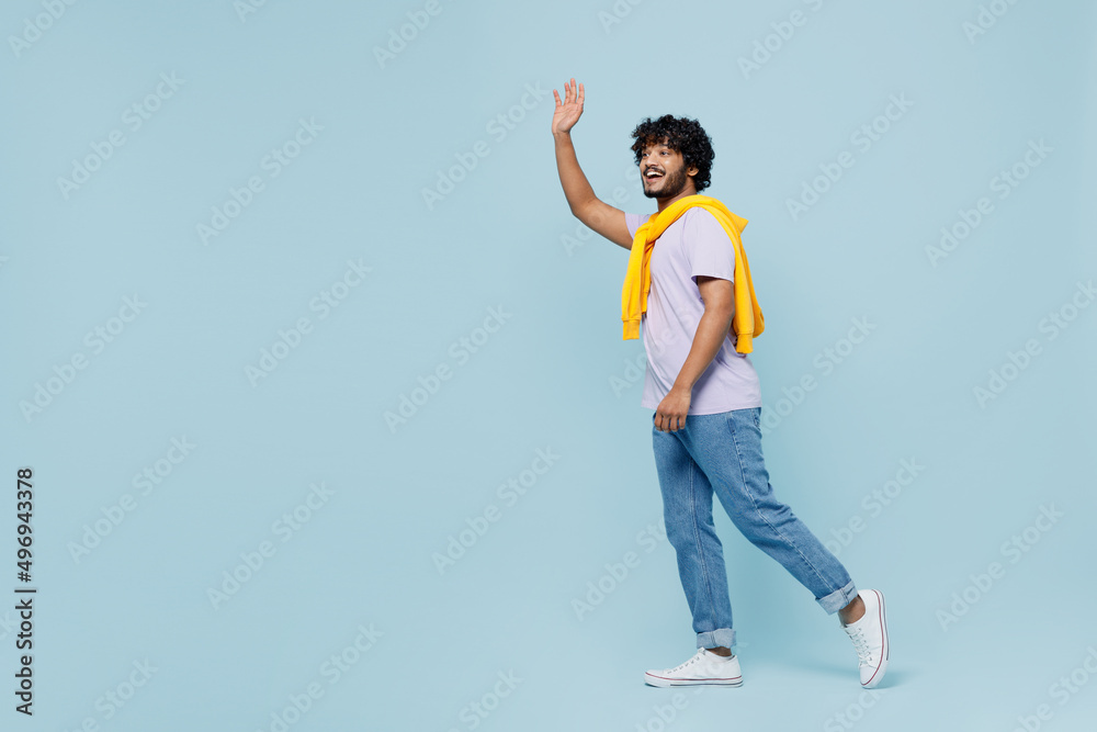 Full size body length side view young bearded Indian man 20s years old wears white t-shirt go meet greet waving hand as notices someone isolated on plain pastel light blue background studio portrait. Full size body length side view young bearded Indian man 20s years old wears white t-shirt go meet greet waving hand as notices someone isolated on plain pastel light blue background studio portrait.