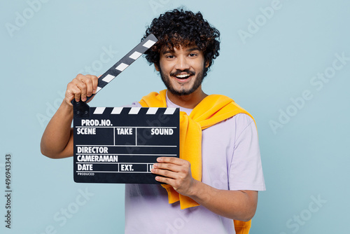 Cheerful happy young bearded Indian man 20s years old wears white t-shirt holding classic black film making clapperboard looking camera isolated on plain pastel light blue background studio portrait.