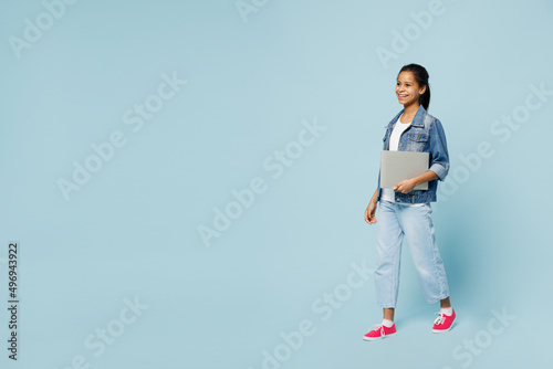 Full body fun little kid teen girl of African American ethnicity 12-13 years old in denim jacket hold laptop pc computer walk going isolated on pastel plain light blue background. Childhood concept.