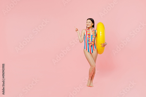 Full body young woman of Asian ethnicity in striped swimsuit hawaii lei hold inflatable ring point finger aside on workspace isolated on plain pink background Summer vacation sea rest sun tan concept