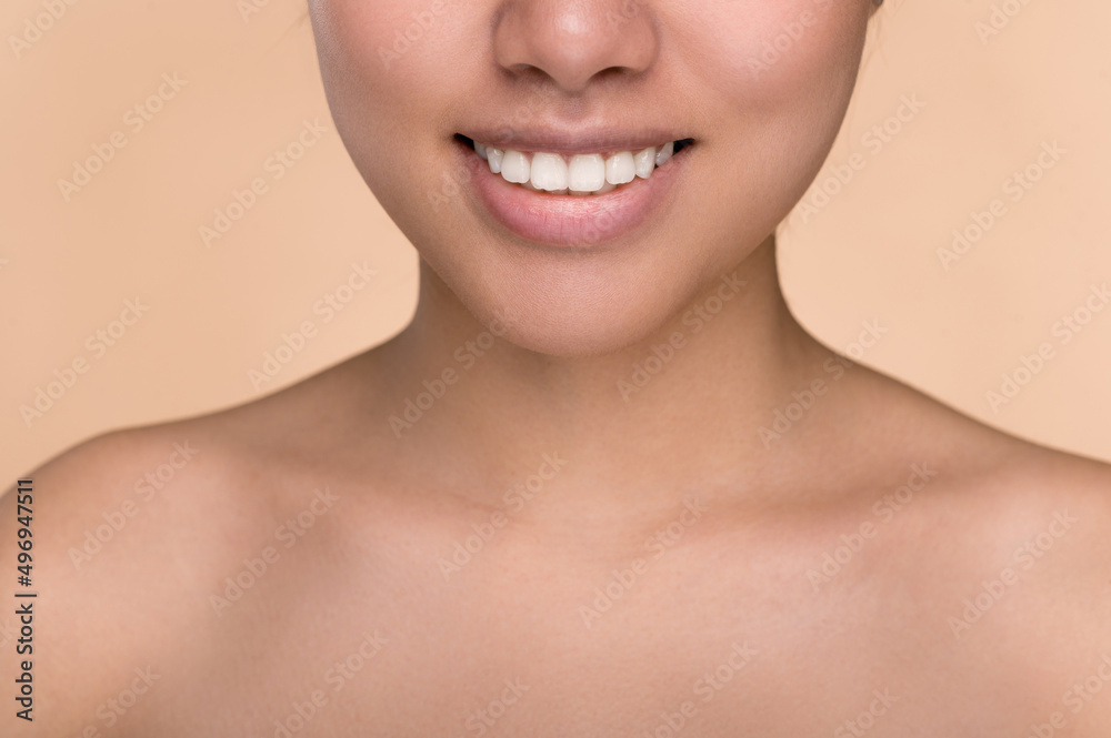 Half face of Asian girl. Chinese young woman with perfect skin demonstrates her smile, perfect white well-groomed teeth, stands on an isolated beige background with naked shoulders. Dentistry concept