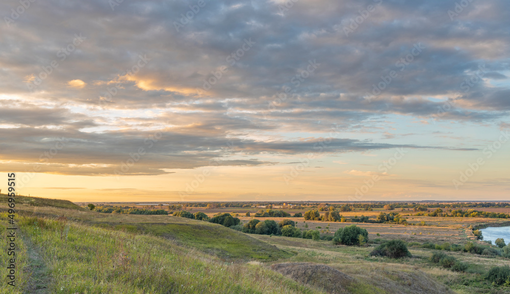 Sunny summer landscape with a river. Nice panoramic view. Green hills, fields and meadows. Dramatic sky over the horizon. Calm. Warm sunlight. Colorful clouds in the blue sky.