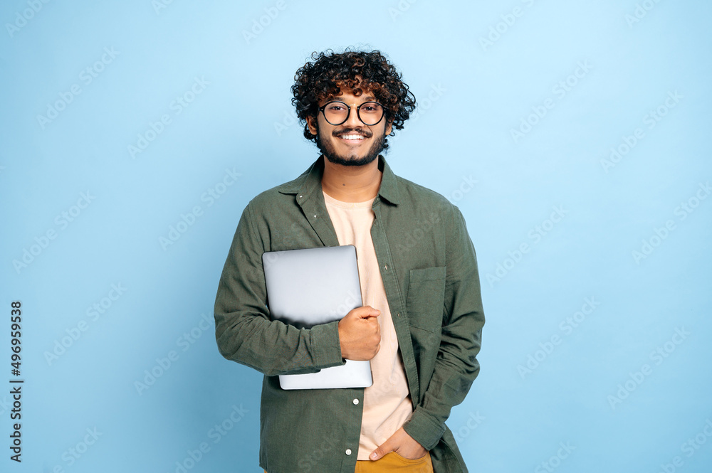 Smart handsome positive indian or arabian guy, with glasses, in casual wear, student or freelancer, holding a laptop in hand, standing on isolated blue background, looking at camera, smiling friendly - obrazy, fototapety, plakaty 