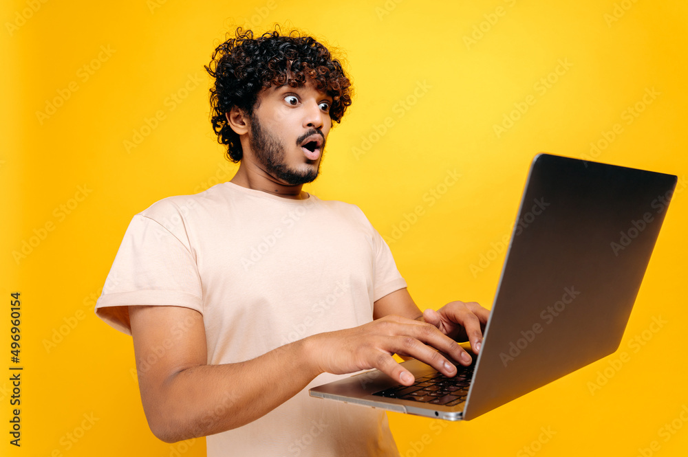 Excited shocked indian or arabian curly-haired guy, typing on a laptop, looking in surprise at the screen, saw unexpected news, victory, profit, stands on isolated orange background. Emotional face Excited shocked indian or arabian curly-haired guy, typing on a laptop, looking in surprise at the screen, saw unexpected news, victory, profit, stands on isolated orange background. Emotional face
